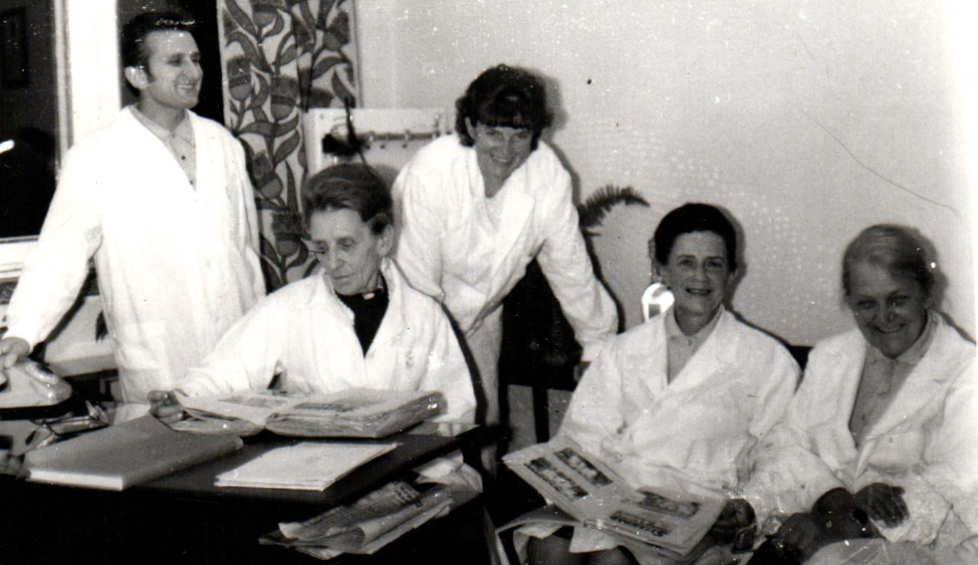 Jeden mężczyzna i cztery kobiety w fartuchach. Oglądają albumy ze zdjęciami, śmieją się.